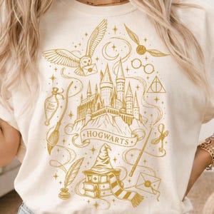 May include: A cream-colored t-shirt featuring a gold-toned graphic print of a castle, an owl, a broomstick, a potion bottle labeled Felix Felicis, a sorting hat on books, a wand, a key, a snitch, glasses, and the text HOGWARTS on a banner.