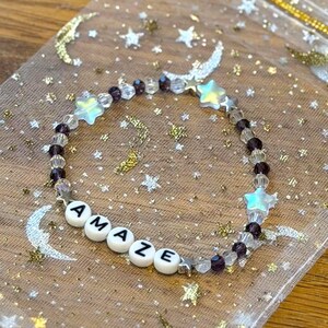 May include: A beaded bracelet with the word "AMAZE" spelled out in white letter beads. The bracelet features clear and purple beads, along with star-shaped and clear beads. It rests on a sheer, gold-speckled bag with moon and star designs.