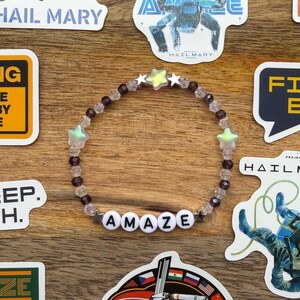 May include: A collection of stickers and a beaded bracelet with the word "AMAZE". The stickers feature space-themed designs, including a spaceship, a robot, and text that reads "Project Hail Mary". The bracelet has star-shaped beads.