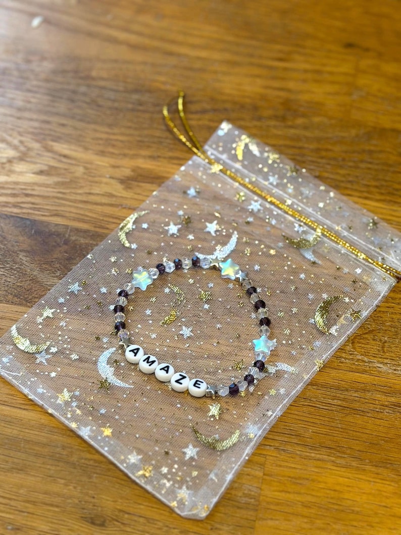 May include: A beaded bracelet with the word "AMAZE" spelled out in white letter beads. The bracelet features clear and purple beads, along with star-shaped and clear beads. It rests on a sheer, gold-speckled bag with moon and star designs.