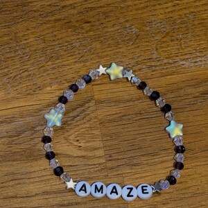 May include: A beaded bracelet with the word "AMAZE" spelled out in white letter beads. The bracelet features clear and purple beads, along with iridescent star-shaped beads.
