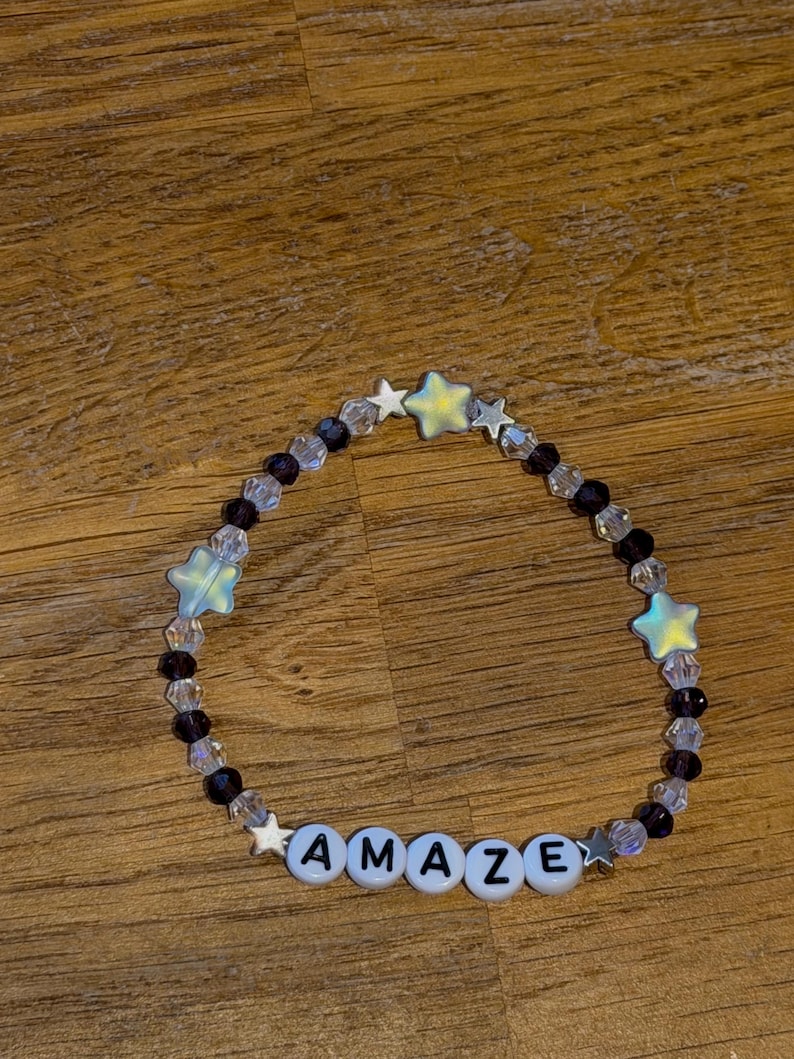 May include: A beaded bracelet with the word "AMAZE" spelled out in white letter beads. The bracelet features clear and purple beads, along with iridescent star-shaped beads.