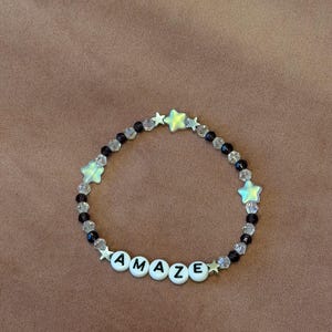 May include: A beaded bracelet with the word "AMAZE" spelled out in white letter beads. The bracelet features clear and black beads, along with star-shaped beads in a light green color. The bracelet is on a brown surface.