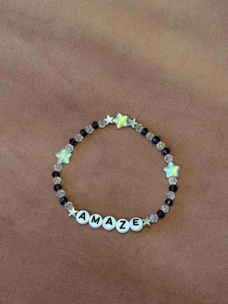 May include: A beaded bracelet with the word "AMAZE" spelled out in white letter beads. The bracelet features clear and black beads, along with star-shaped beads in a light green color. The bracelet is on a brown surface.