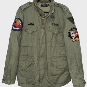 Puede incluir: Chaqueta de estilo militar verde oliva con múltiples bolsillos, parches y el texto "US-RL". La chaqueta tiene cierre de cremallera y cintura ajustable. Los parches incluyen un emblema alado y una calavera con gafas.