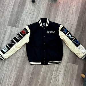 May include: A black varsity jacket with cream-colored sleeves. The jacket features patches on the sleeves and the word "Human" embroidered on the chest. White stripes accent the collar, cuffs, and hem.
