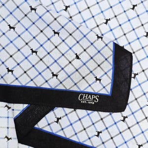 May include: A white pocket square featuring a blue and black plaid pattern with small black dog silhouettes throughout. The fabric has a wide black border with a thin blue inner stripe and the white text CHAPS EST. 1978 printed in the corner.