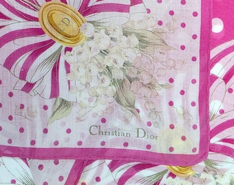 May include: A pink and white silk scarf featuring a floral and bow design. The scarf has a polka dot pattern and a border. The text "Christian Dior" is printed on the scarf.