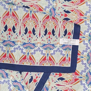 May include: A square fabric scarf featuring a repeating floral pattern in red, blue, and cream with a solid navy blue border. A white rectangular tag is attached to the edge, printed with the text INTERMODE Using LIBERTY ART FABRICS.