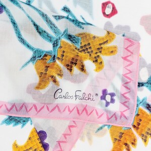 May include: A white scarf featuring a colorful floral pattern with yellow, teal, and purple elements. The fabric has a pink border with a zigzag design and the text Carlos Falchi printed in black script near the center of the folded edge.