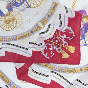 May include: A folded white and red scarf with a repeating pattern of horse-drawn carriages, gold accents, and the word "CELINE". The scarf features a detailed design with purple, gold, and black elements.