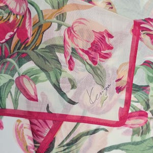 May include: A white scarf featuring a vibrant floral pattern of pink and red tulips with green leaves and stems. The fabric has a semi-sheer texture and is finished with a solid pink border. The word Kenzo is printed in a cursive script on the fabric.