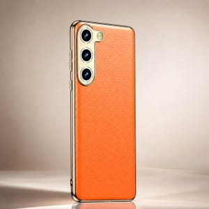 Op de afbeelding: Oranje leren telefoonhoesje met een goudkleurig frame. De hoes heeft drie cameralenzen en een flitser. De telefoonhoes is ontworpen om de telefoon te beschermen tegen krassen en beschadigingen.