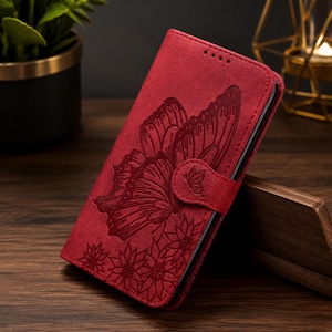May include: A red wallet-style phone case featuring an embossed butterfly and floral pattern on the front cover. The case includes a magnetic closure tab with a small butterfly detail and four small circular cutouts at the top edge.