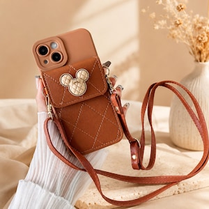 Op de afbeelding: Een bruine telefoonhoes met een doorgestikte textuur en een bijpassende schouderriem. De hoes heeft een klepzak aan de achterkant, versierd met een beige, doorgestikt Mickey Mouse-hoofd embleem. De camera-uitsparing heeft een bruine rand.