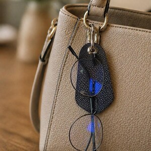 Peut inclure: Un sac à main en cuir beige avec un porte-lunettes noir attaché. Le support présente un motif texturé et contient une paire de lunettes à monture ronde. Le sac a une quincaillerie dorée.