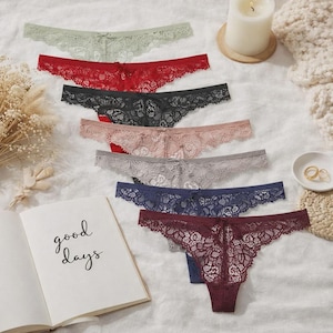Könnte beinhalten: Eine Auswahl von sieben Spitzen-Tanga-Unterhosen in verschiedenen Farben, darunter Grün, Rot, Schwarz, Rosa, Grau, Marineblau und Burgunderrot. Jeder Tanga hat ein zartes Spitzendesign und eine kleine Schleife. Ein Buch mit den Worten "good days" ist ebenfalls zu sehen.