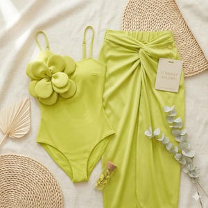 Puede incluir: Un traje de baño verde lima con un gran detalle floral y tirantes finos se combina con una falda a juego. La falda tiene un detalle de nudo en la cintura. Una pequeña tarjeta dice "VERDANT STUDIO".