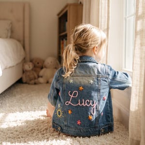 May include: A blue denim jacket with the name "Lucy" embroidered in pink script on the back. The jacket is decorated with flower and sun patches. The child is sitting on a white rug.