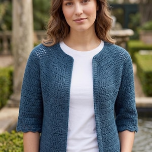 May include: A teal blue, open-front crochet cardigan with three-quarter sleeves. The cardigan features a scalloped edge and a textured stitch pattern. The person is wearing a white t-shirt underneath. The background is a garden setting.