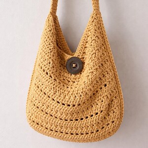 May include: Mustard yellow crochet shoulder bag with a large, round, dark brown button closure. The bag has a wide strap and a textured, open weave design. The bag is hanging against a plain white background.