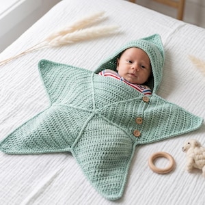 May include: A light green crocheted star-shaped baby swaddle blanket with a hood. The swaddle features three small wooden buttons along one side and a textured stitch pattern. The baby inside wears a red, white, and blue striped shirt.