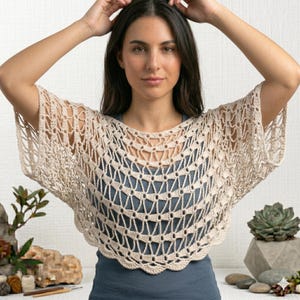 Peut inclure: Un poncho en crochet beige à mailles ajourées, avec une bordure festonnée. Le poncho a une encolure ronde et une coupe ample et fluide. Le motif au crochet crée un aspect de filet. Le vêtement est porté sur un haut bleu.