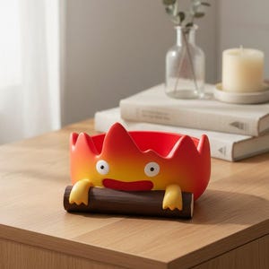 May include: A red and yellow decorative bowl shaped like a cartoon fire spirit, holding a brown log. The bowl is on a wooden surface, with a candle, books, and a vase with greenery in the background.