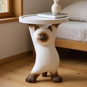 May include: A whimsical white cat-shaped side table with a round top. The cat figure has brown accents on its paws, ears, and face, and appears to be holding up the tabletop. A small vase and a book rest on the table's surface.