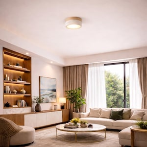 May include: A modern living room with a neutral color palette. A round, white and gold coffee table sits in front of a cream-colored sofa with several pillows. A wooden bookshelf and a painting are on the wall. A ceiling light fixture with a wooden trim is in the center.