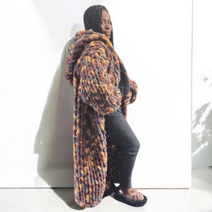 May include: A long, hooded cardigan in a chunky knit pattern with shades of brown, orange, and purple. The cardigan is worn by a person standing in front of a white wall.