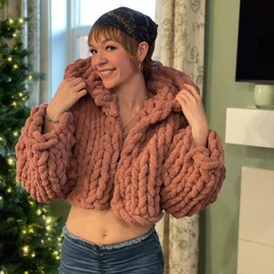 Peut inclure: Un pull rose en tricot court avec une capuche. Le pull est fait de fil épais et a une coupe ample. Le mannequin porte un jean bleu.