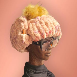 May include: A pink and purple fuzzy hat with a yellow pom pom on top. The hat is worn on a mannequin with black framed glasses.