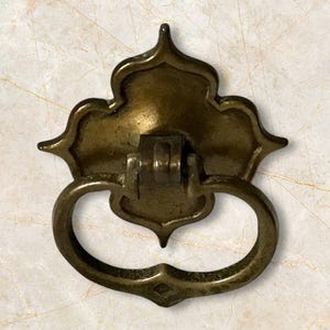 May include: Antique brass drawer pull with a decorative backplate and a curved handle. The backplate has an ornate, symmetrical design with pointed edges. The handle is a rounded shape with a diamond-shaped detail at the bottom.