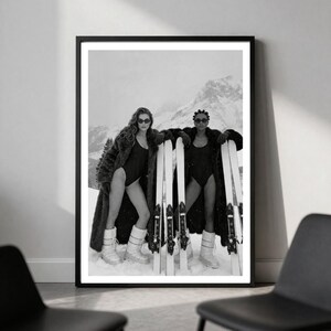May include: A black and white framed wall art print featuring two individuals in swimsuits, fur coats, and snow boots, each holding a pair of skis. They are wearing dark sunglasses and standing in a snowy mountain landscape with peaks in the background.