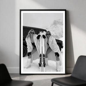May include: A black and white framed wall art print depicting a person wearing a long faux fur coat, dark sunglasses, and gloves, standing in the snow next to a pair of skis and a vehicle. The scene features a mountainous landscape in the background.