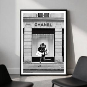 Puede incluir: Impresi&oacute;n en blanco y negro enmarcada que muestra a una persona corriendo con bolsas de compras frente a una tienda Chanel. El nombre de la tienda es visible sobre la entrada. Una imagen elegante que captura la esencia del lujo y la moda.