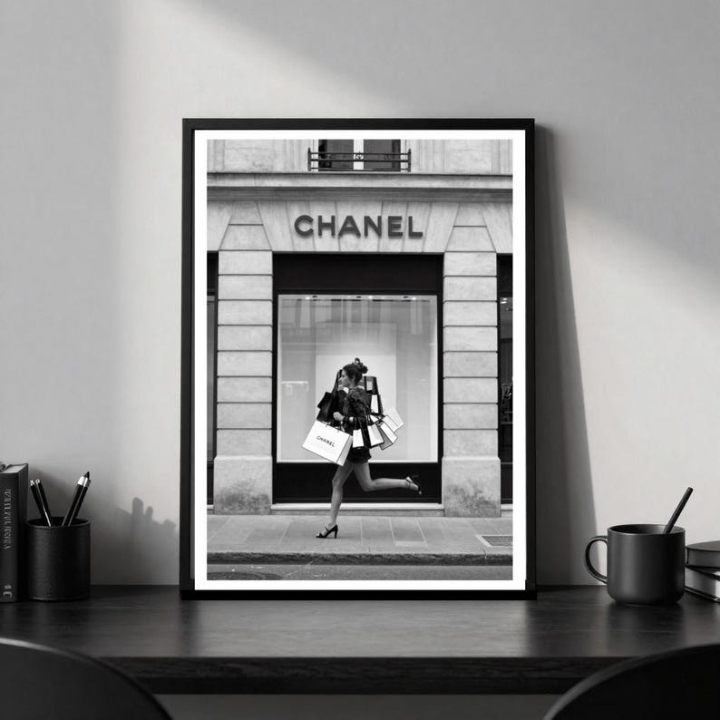 Puede incluir: Impresi&oacute;n en blanco y negro enmarcada que muestra a una persona corriendo con bolsas de compras frente a una tienda Chanel. El nombre de la tienda es visible sobre la entrada. La imagen captura la compra de lujo y moda.