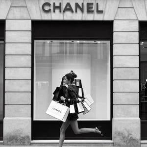 Puede incluir: Imagen en blanco y negro de una mujer corriendo con varias bolsas de compras frente a una tienda Chanel. Las bolsas son blancas con el logo de Chanel. La tienda tiene un gran escaparate y el logo de Chanel encima de la entrada.