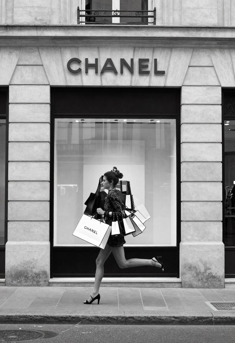 Puede incluir: Imagen en blanco y negro de una mujer corriendo con varias bolsas de compras frente a una tienda Chanel. Las bolsas son blancas con el logo de Chanel. La tienda tiene un gran escaparate y el logo de Chanel encima de la entrada.