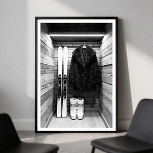May include: A black and white framed print depicting a wooden closet interior. Inside, a dark faux fur coat hangs on a hanger between two tall skis. Below the coat, a pair of white winter boots with the brand name Chanel are placed on the floor.