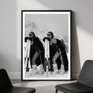 May include: A black and white framed wall art print featuring two identical figures wearing dark swimsuits, long fur coats, sunglasses, and white snow boots. They are standing in a snowy mountain landscape, each leaning against a pair of skis.