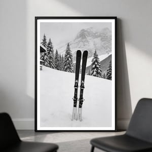 May include: A black and white framed wall art print depicting a pair of black skis standing upright in deep snow. The background features a snowy mountain slope covered with evergreen trees under a cloudy sky, captured in a monochromatic style.