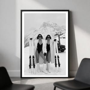 May include: A black and white framed wall art print featuring two individuals standing in snow, wearing dark swimsuits, white faux fur coats, and white moon boots. Each person holds a pair of skis. A ski lift and a dark SUV are visible in the background.