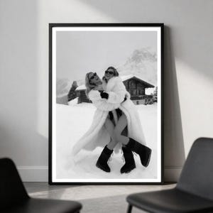 May include: A black and white framed wall art print depicting two individuals wearing long, fluffy white coats, dark sunglasses, and black boots while embracing in a snowy mountain landscape with a wooden cabin visible in the background.