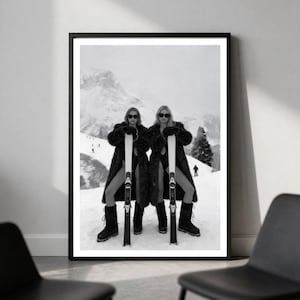 May include: A black and white framed wall art print depicting two individuals standing in a snowy mountain landscape. Both figures wear dark fur coats, dark sunglasses, and dark boots while holding upright skis. The scene features a vast, snow-covered slope with distant mountain peaks and small figures in the background.