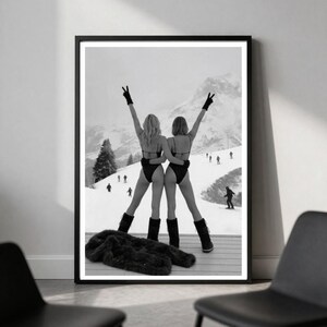 May include: A black and white framed art print depicting two individuals in black swimsuits, gloves, and boots standing on a balcony overlooking a snowy mountain ski slope. The individuals have their arms raised in a V-shape against a winter landscape.