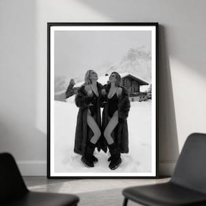May include: A black and white framed wall art print depicting two individuals wearing dark bodysuits and long fur coats, standing in a snowy mountain landscape with a wooden cabin in the background, each holding a stemmed glass in their hand.