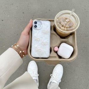 Puede incluir: Una funda de teléfono blanca con detalles de lazo de perlas y un borde de pedrería. La funda está en un portavasos de cartón con un café helado, una funda de auriculares inalámbricos blanca y un lápiz labial rosa. La persona lleva zapatillas blancas y pulseras doradas.