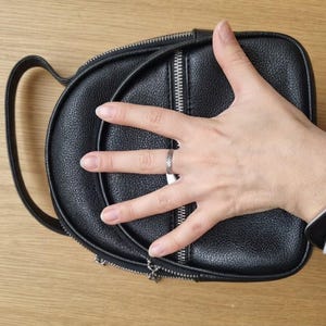 May include: A small black backpack with a textured, pebbled finish. The bag features a rounded top handle, a vertical zippered front pocket with silver-toned hardware, and a main compartment zipper closure. The design is compact and structured.
